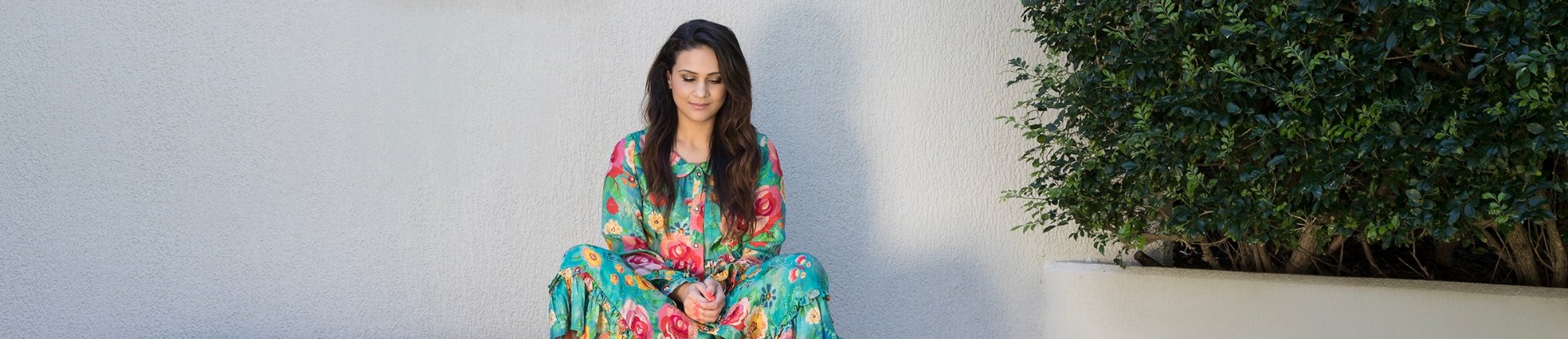 Esha Oberoi sitting cross-legged against a pale wall in a green floral dress, eyes lowered