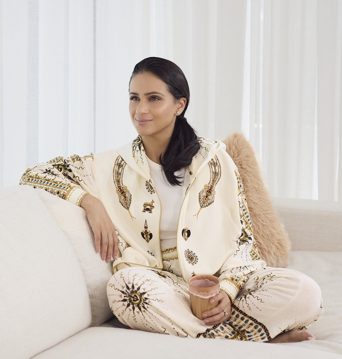 Esha Oberoi sitting cross-legged on a cream sofa, holding a cup