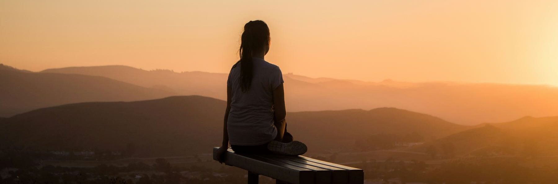 A woman sitting on a bench overlooking hills at sunset