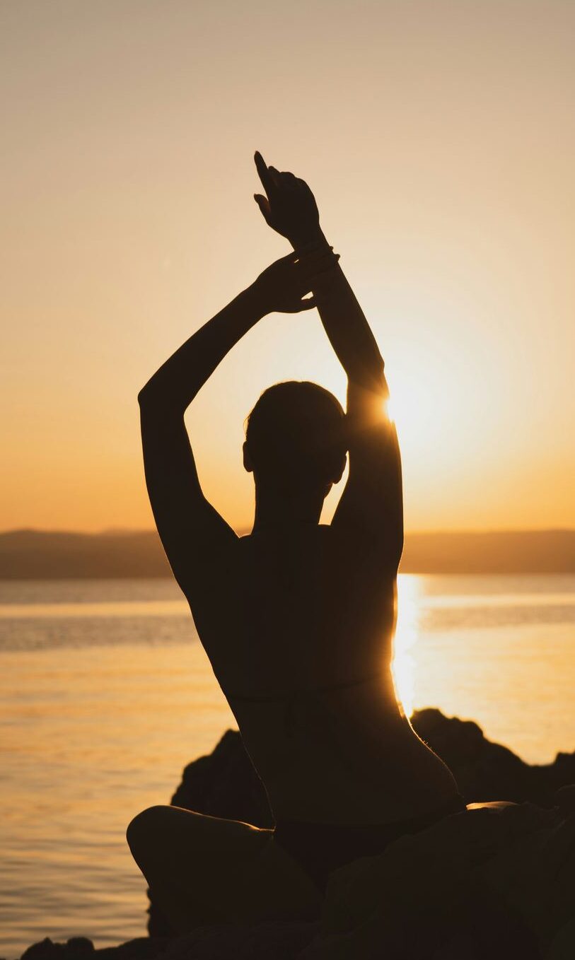 Silhouette of a woman with arms raised beside the sea at sunset